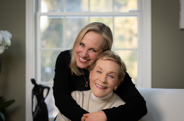 A younger woman stands behind and hugs an older woman seated on a white sofa. Both are smiling in a bright room with large windows.