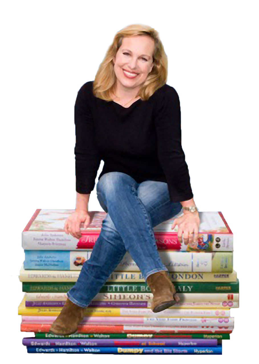 A woman with blonde hair, wearing a black top and blue jeans, sits smiling on a stack of large, colorful books against a white background.
