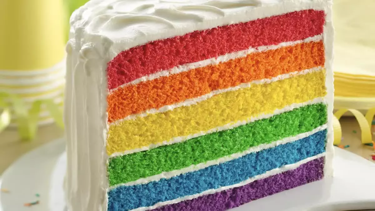 A slice of cake with six layers in rainbow colors, covered with white frosting, sits on a white plate.