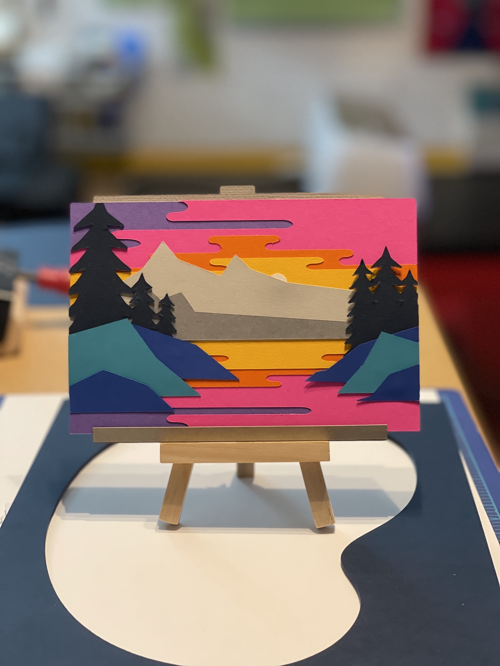 A colorful paper art landscape of mountains, trees, and a lake at sunset, displayed on a small wooden easel indoors.