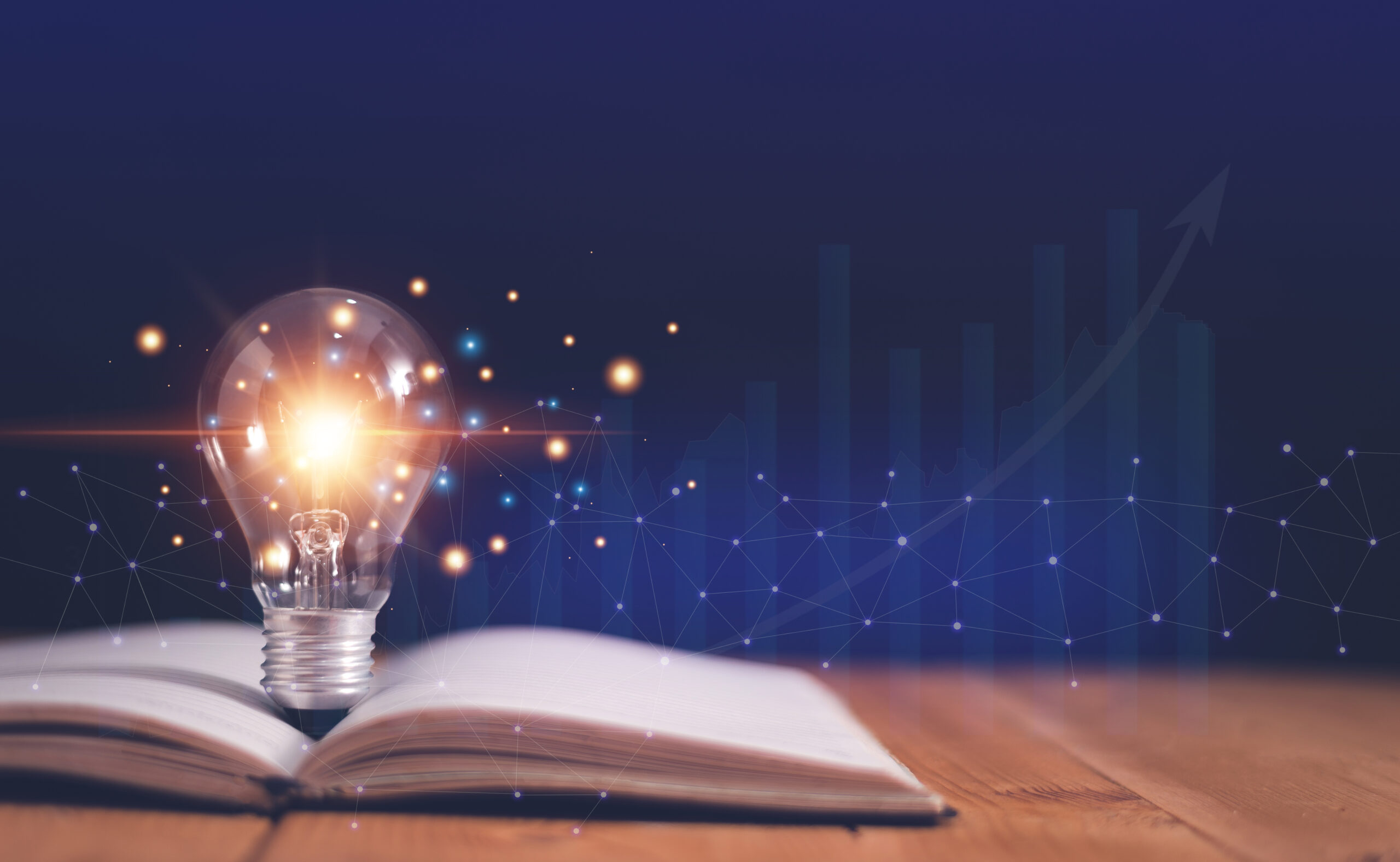 A glowing light bulb sits on an open book on a wooden table, with digital graphics and rising bar charts in the background.