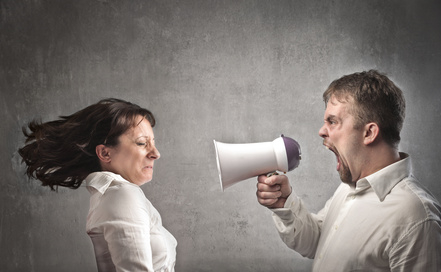 A man shouts into a megaphone aimed at a woman's face; she reacts by flinching away. Both wear white shirts and stand against a gray background.